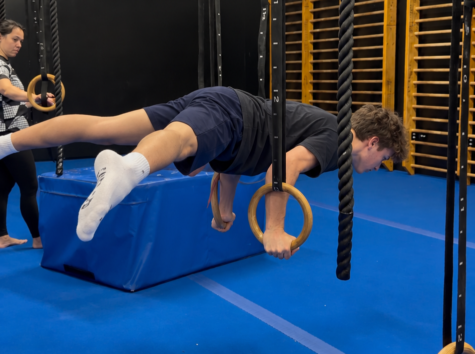 Teenager practising a controlled ring strength position while a coach supervises in the Falsegrip gym.